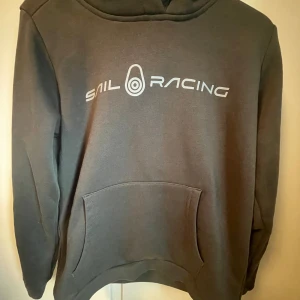 Svart/grå hoodie från Sail Racing - Svart/grå hoodie från Sail Racing med stor logga framtill. Klassisk känguruficka och huva. Perfekt för dig som gillar en enkel och stilren look. Mjuk insida och långärmad modell. Säljer för den är för liten köpte för mindre än en månadsedan, använd en gång 