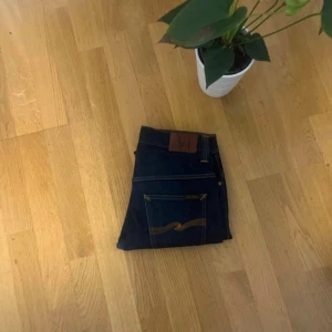 Mörkblå Nudie Jeans straight fit - Beskrivning: Säljer ett par äkta Nudie Jeans i mörkblå denim. Klassiska med den ikoniska vågiga sömmen på bakfickorna och läderpatch i midjan. Byxorna är i fint skick, knappt använda och utan skador eller fläckar. Passar perfekt både till vardags och mer dressat.  Nypris ligger runt 1 500–1 800 kr.  ✅ Äkta Nudie Jeans ✅ Snygg mörk denim som åldras fint ✅ Bra kvalitet och komfort  Finns att hämta i [din stad] eller skickas mot fraktkostnad.
