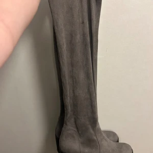 Grå mockastövlar med klack - Snygga höga stövlar i grå mocka med rund tå och kraftig klack. Stövlarna har en stilren design och passar perfekt till både jeans och kjol. Materialet är mjukt och ger en lyxig känsla. Klacken ger extra höjd utan att tumma på komforten.