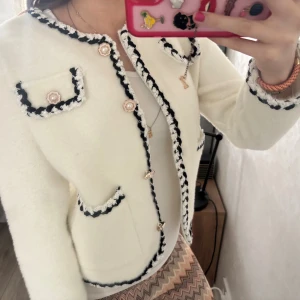 Vit cardigan jacka med pärlknappar - Superfin vit cardigan jacka med svarta och vita virkade kanter och dekorativa pärlknappar. Jackan har fyra fickor framtill och är långärmad. Den är mjuk och har en klassisk, elegant look som passar perfekt till jeans eller kjol.