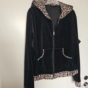 Svart velourhoodie med leoparddetaljer - Storlek M . Jätte fint skick.  Svart hoodie i mjuk velour från Triumph med leopardmönstrade detaljer på huva, ärmslut, fickor och nederkant. Dragkedja framtill och kängurufickor. Perfekt statement-plagg för dig som gillar djurmönster och comfy vibes. 