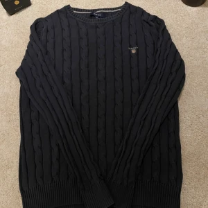 Mörkblå kabelstickad tröja från GANT - Säljer en mörkblå kabelstickad tröja från GANT i storlek S. Tröjan har rund halsringning, ribbade muddar och GANT-logga broderad på bröstet. Perfekt för dig som gillar klassisk och stilren look.