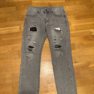 Grå slitna jeans med hål - Säljer ett par grå jeans med flera slitningar och hål framtill för en riktigt edgy look. Byxorna har klassisk femficksdesign och raka ben. Materialet är 83% bomull och 17% polyester, vilket gör dem både mjuka och slitstarka. Perfekta för dig som gillar streetstyle.