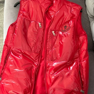 Röd dunväst från Moncler - Snygg röd dunväst från Moncler med glansig finish och klassisk logga på bröstet. Västen har flera dragkedjefickor framtill och hög krage. Perfekt för lager-på-lager och ger en sportig vibe. Fodrad insida med coolt Moncler-tryck.