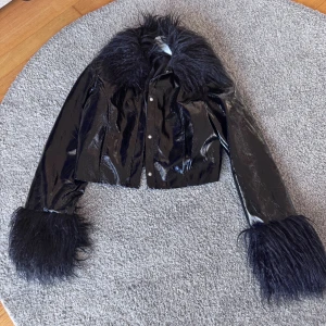 Svart pälskantad jacka med glans - Säljer en svart, glansig jacka med stora pälskanter vid krage och ärmslut. Jackan är croppad och har tryckknappar framtill. Materialet är syntet med pälsdetaljer som ger en riktigt statement-look.