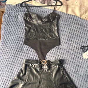 Svart skinnkjol och en body från Zara - Supersnygg svart kort skinnkjol från Zara med markerade sömmar och ett brett bälte med guldig detalj i midjan. Med den kommer även en body man kan matcha med. Pris kan diskuteras vid snabb köp 