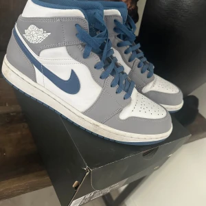 Nike Air Jordan 1 Mid Cement Grey/Blue - Nike Air Jordan 1 Mid i cementgrått, vitt och blått. Klassisk high-top siluett med blå snörning, blå swoosh och blå yttersula. Ovandel i skinn och syntet, perforerad tåbox och ikonisk Air Jordan-logga på sidan. Perfekta för dig som vill sticka ut med stil.