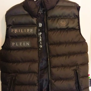 Svart dunväst från Philipp Plein - Snygg svart dunväst från Philipp Plein med quiltad design och logotyp på bröstet. Västen har dragkedja framtill, två fickor med dragkedja och tydliga logodetaljer. Perfekt för lager-på-lager och streetstyle.