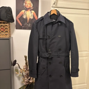Mörkblå trenchcoat från Zara - Snygg mörkblå trenchcoat från Zara med klassisk dubbelknäppning, axelklaffar och bälte i midjan. Jackan har långa ärmar, stora fickor och stilren krage. Perfekt för dig som vill ha en tidlös och trendig look.
