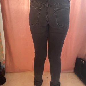 Mörkgrå skinny jeans i storlek S - Säljer ett par mörkgrå skinny jeans i storlek S. Jeansen har hög midja, tjejen som har på sig dom e 149/150 och dom e för långa klassiska fem fickor och smal passform hela vägen ner. Materialet är stretchigt och bekvämt, perfekt för dig som gillar en tight look. Snygga detaljer med svarta sömmar och knappstängning.