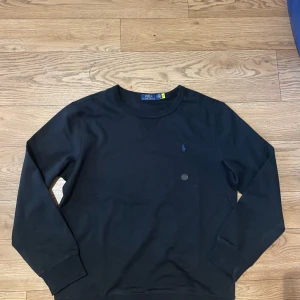Svart tröja från Polo Ralph Lauren - Svart långärmad tröja från Polo Ralph Lauren med klassisk rund halsringning och broderad logga på bröstet. Tillverkad i mjuk bomull för en skön och avslappnad känsla. Perfekt basic-plagg med stilren look. 