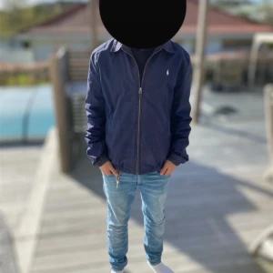 Mörkblå vindjacka från Ralph Lauren - Snygg mörkblå vindjacka från Ralph Lauren med klassisk krage och dragkedja framtill. Jackan har elastiska muddar vid ärmslut och nederkant samt den ikoniska lilla loggan broderad på bröstet. Perfekt för dig som gillar stilren och enkel design.