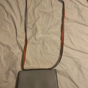 Ljusgrå axelväska i skinn - Stilren ljusgrå axelväska i slätt skinn med justerbart axelband i kontrasterande brunt. Väskan har rektangulär form, magnetlås och flera fack inuti för smart organisering. Perfekt för dig som gillar minimalistisk och clean design.