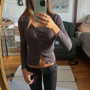 Mörkgrå ribbad topp med knappar - Säljer en mörkgrå ribbad topp med lång ärm och snygg v-ringning. Toppen har en croppad passform och knappar längs hela framsidan. Materialet är stretchigt och sitter tight mot kroppen, perfekt för en trendig look.