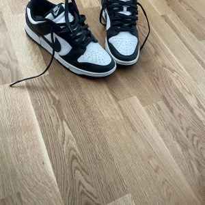 Nike Dunk Low svart/vit sneakers - Nike Dunk Low sneakers i svart och vitt med klassisk Swoosh på sidan. Skorna har snörning, perforerad tå och platt sula. Ovandelen är i skinn och syntet, vilket ger en clean och tidlös look. Perfekta för dig som gillar streetstyle och vill ha en ikonisk sneaker i garderoben.