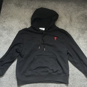Svart hoodie från Ami med rött hjärta - Svart hoodie från Ami Paris huva, snörning och känguruficka. På bröstet finns Ami:s ikoniska röda hjärta med A-broderi. Tröjan är i mjuk bomull och har ribbade muddar vid ärmslut och nederkant.