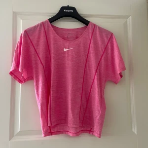 Rosa Nike Dri-Fit t-shirt - Sportig rosa t-shirt från Nike med Dri-Fit-teknologi. T-shirten har korta ärmar, rund halsringning och en vit Nike-logga på bröstet. Materialet är lätt och andas, perfekt för träning. Snygga sömdetaljer framtill ger en modern look.