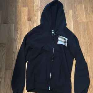 Svart hoodie med dragkedja och Burberry-detalj - Svart hoodie från Burberry med klassiskt rutigt tyg på bröstet. Tröjan har huva, dragkedja framtill och två fickor. Tillverkad i mjukt bomullsmaterial och har långa ärmar. Perfekt för en chill och stilren look.