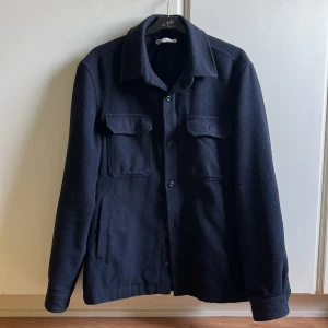 Mörkblå overshirt i ull med fickor - Stilren mörkblå overshirt i ull med klassisk krage och knäppning framtill. Två stora bröstfickor med lock och två sidofickor ger en clean och praktisk look. Perfekt lager-på-lager-plagg för kyligare dagar. Väldigt bra skick och sparsamt använd, skriv för fler bilder!
