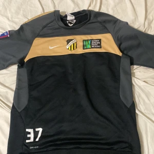 BK Häcken långärmad Nike fotbollströja - BK Häcken långärmad fotbollströja från Nike i svart och guld med klubbmärke, sponsortryck och nummer 37. Tillverkad i Dri-Fit-material som andas. Stadium-logga på ryggen och Stryktipset-märke på ärmen. Perfekt för dig som gillar fotboll och vill sticka ut på planen.