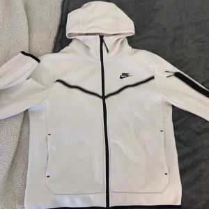 Vit Nike hoodie med svarta detaljer - Säljer en vit hoodie från Nike med svarta detaljer och dragkedja hela vägen. Tröjan har huva, lång ärm och en snygg svart Nike-logga på bröstet. Praktiska fickor och en extra ficka med dragkedja på ärmen. Perfekt för dig som gillar sportig stil.