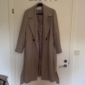 Beige/grå lång kappa från Stradivarius - Jätte fin beige/grå lång kappa från stradivarius perfekt inför hösten. Använd fåtal gånger den värkligen färgen framkommer mest på bilderna nära 