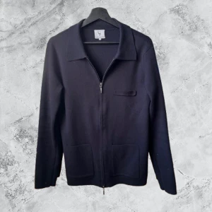 Svart kofta från Urban Pioneers - Stilren svart kofta från Urban Pioneers i storlek S. Koftan har dragkedja framtill, klassisk krage och två praktiska fickor. Materialet är mjukt och stickat, perfekt för lager-på-lager. Enkel design som funkar till många olika looks.