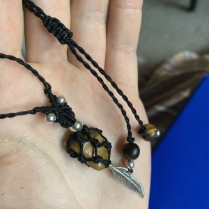Kristall halsband - Makramé halsband med äkta tigeröga-stenar. Stenen är 1,8cm X 1,4cm och pärlorna i ändan 8mm. Med en tillhörande fjäder under stenen i rostfritt stål. Halsbandet går att ställa in till den längd du önskar. Halsbandet tål vatten.