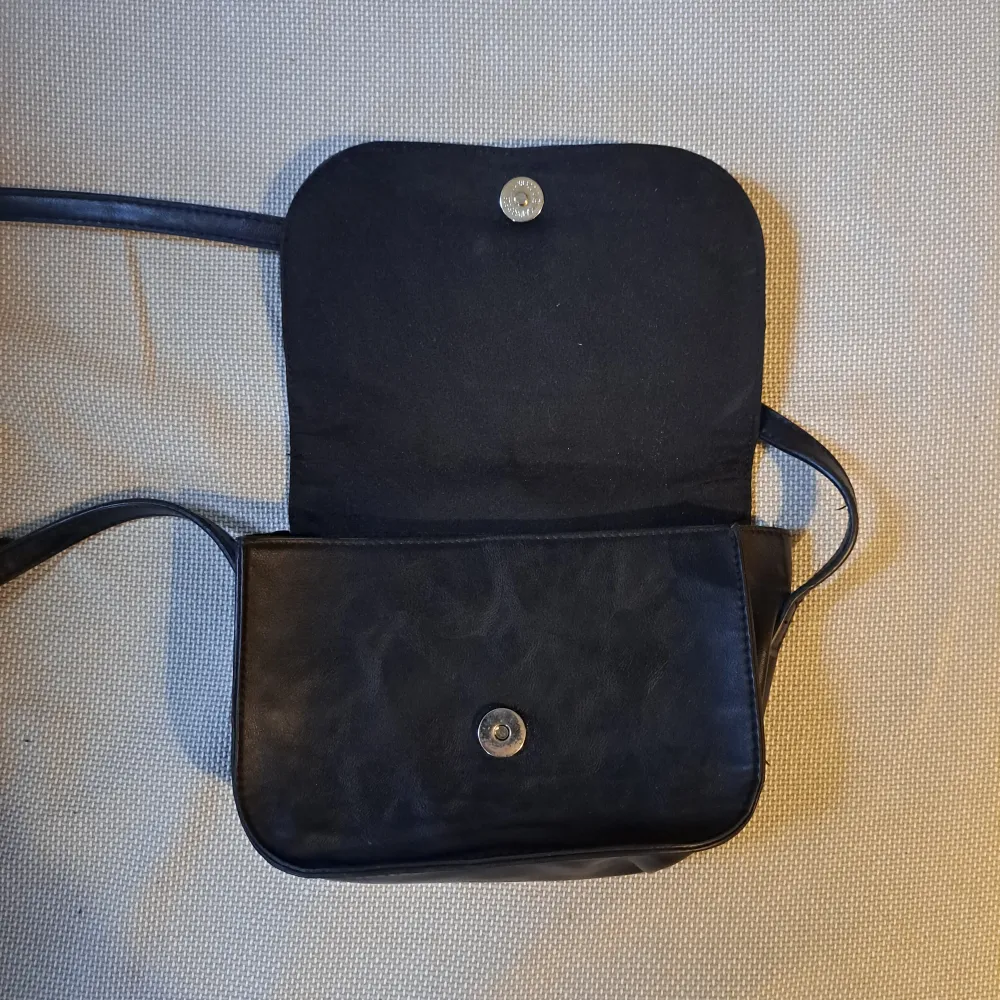 En svart axelremsväska från Even&odd. Väskan är överlag i bra skick förutom missfärgningen på den undre knappen. Väskor.