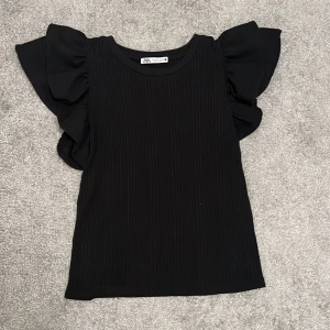 Svart T-shirt med volang axlar - Svart T-shirt med volang axlar, använd endast 1 gång, inga skador 