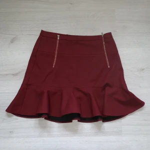 Vinröd lågmidjad kjol - Vinröd lågmidjad kjol med dragkedjor framtill och volang. Kjolen är i bra skick, vet ej var den är ifrån. Storlek M men är mer som XS/S.