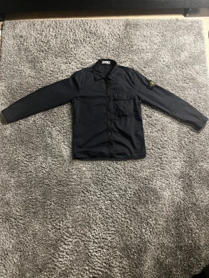 Stone island overshirt  - En perfekt sommar jacka till salu som vi på Blanco har löst till er. Det är en marinblå färgad overshirt i ett utsökt skick. Vi på Blanco säljer denna för 1650kr då ny pris på denna är 4,500kr. Tveka inte på höra av er! Mvh Blanco