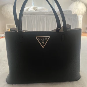 Guess handväska - Väska använd 1 gång, nyskick. Köpt i Stockholm på Åhléns för 2000kr! Har ett fack i mitten som går att stänga med dragkedja. Väskan kan stängas med en magnetisk knapp. Jättefin väska som passar till allt!  Väskan är 36x27x15cm!