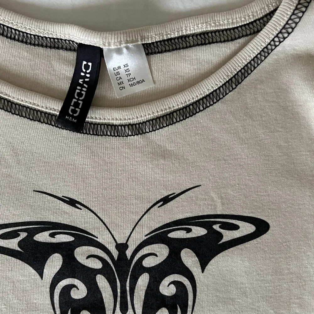 Jättesöt beige croppad t-shirt från H&M med tryck av en fjäril på framsidan. Aldrig använd, nyskick. Storlek XS men passar även S💕🫶. T-shirts.