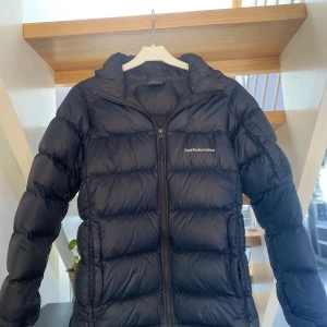 Peak performance jr frost Down Hood jacket  - Säljer en svart peak performance jr frost Down Hood jacket i storlek 160!  logga är broderad och placerad på bröstet och på ärmen. Jackan har två fickor framtill som stängs med dragkedja. Säljes pga för liten! nypris 1799kr mitt pris 299kr