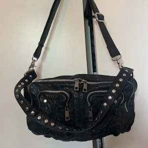 Nunoo väska  - Nyskick, knappt använd.  Nypris: 1399:- Väska i äkta skinn Två axelremmar i olika längd medföljer, den ena med dekorativa nitar. Två fack på insidan varav ett försluts med dragkedja. Tre fack på utsidan som förslut. Bredd: 24,5 cm, Höjd: 15 cm, Djup: 8 cm