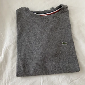 T-shirt från Lacoste i storlek XS - En söt, grå T-shirt från märket Lacoste i storleken XS. Tröjan är i fint skick då den inte kommit till användning. Den är i barnstorleken ”164” och motsvarar   en XS i damstorlekar.
