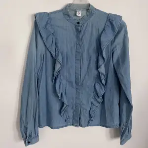 En blus i tvättad denim av ekologisk bomull. Blusen har låg ståkrage, dekorativa volanger och dold knäppning fram.   