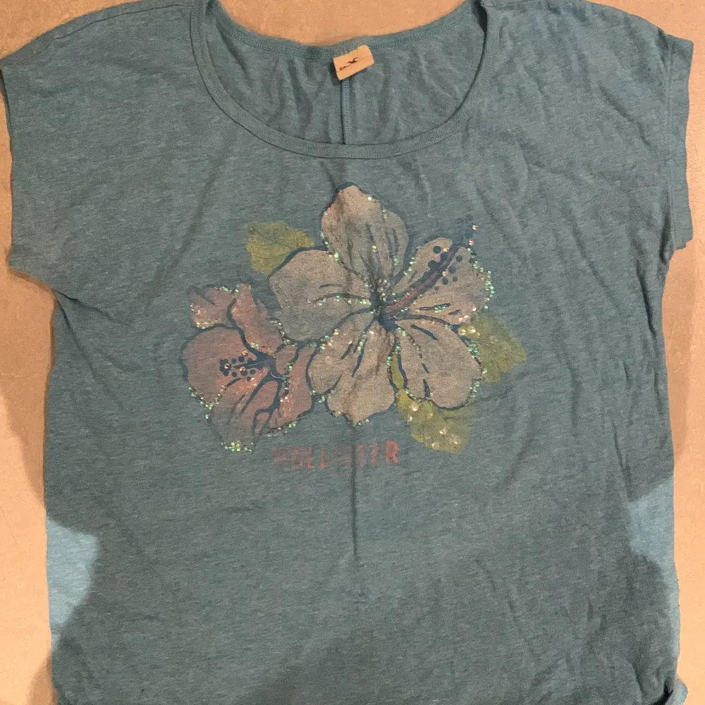 En blå tröja med paljetter med en hibiskus Bloms på. Köpte den secondhand men använder inte den så mycket tyvärr. Den är urringad. Somrig. T-shirts.