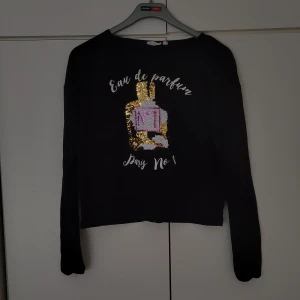 Svart topp med glittertryck - Svart topp med glittertryck från Kappahl i storlek 170. Paljetter med ett tryck på framsidan, se bild. Längden från hals till midja är ca. 44 cm.Armlängden från armhålan är ca. 51 cm. Tunnt, skönt material, som är lite stretch i.