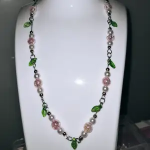 Ett vackert halsband som består av vita och rosa akrylpärlor, roséguld pärlor, gröna blad och guldigt hjärta. 