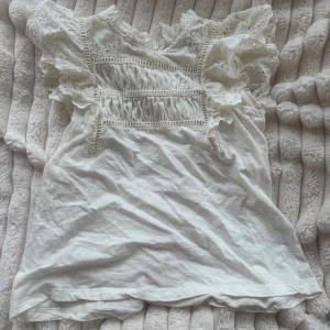 Vit spetsblus från Holly & Whyte - Supersöt vit blus från Holly & Whyte med volangärmar och broderade spetsdetaljer. Blusen har rund hals, romantiska hålbroderier och är tillverkad i mjuk bomull. Perfekt för sommarens alla tillfällen!