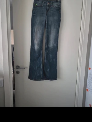 Blå bootcut jeans - Säljer ett par blå bootcut jeans från Booty med snygga slitningar och tvättade detaljer. Jeansen har klassisk femficksdesign, låg midja och är tillverkade i en mix av bomull, polyester och elastan för skön stretch. Perfekta för dig som gillar retrovibbar och markerad siluett. Storleken är 23/30 och säljer då de blivit för små och ska köpa nya.