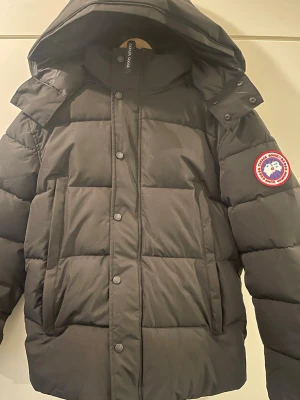 Svart dunjacka från Canada Goose - Säljer en svart dunjacka från Canada Goose med klassisk patch på ärmen. Jackan har huva, stora fickor och stängs med både dragkedja och knappar. Perfekt för kalla vinterdagar och riktigt snygg streetstil.