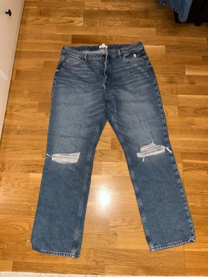 Blå ripped jeans från H&M, strl 48 - Säljer ett par blå jeans från H&M i storlek 48 med raka ben och slitningar över knäna. Jeansen har klassisk femficksdesign och är gjorda i bomull. Perfekta för en avslappnad och trendig look.