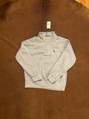 Grå half zip från Polo Ralph Lauren - Snygg grå långärmad tröja från Polo Ralph Lauren med half zip och klassisk broderad logga på bröstet. Tröjan har ribbade muddar och är gjord i mjuk bomullsmix, perfekt för lager-på-lager under höst och vinter.