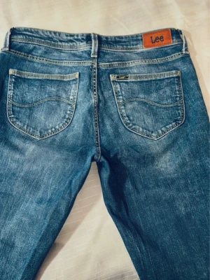 Blå raka jeans från Lee W27/L31 - Säljer ett par klassiska blå jeans från Lee med raka ben och normal passform. Jeansen har fem fickor, snygga slitningar och kontrastsömmar. Perfekta till vardags och funkar till det mesta. Tidlös modell med Lee-lapp bak i midjan.