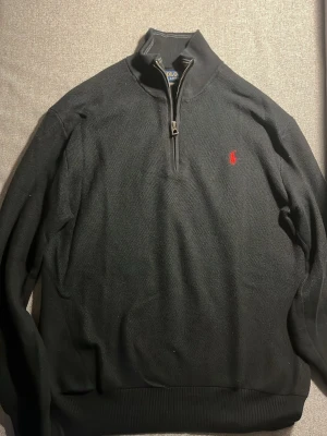 Svart half zip tröja Polo Ralph Lauren - Klassisk svart tröja från Polo Ralph Lauren i mjuk pima bomull. Half zip-dragkedja och röd broderad logga på bröstet. Ribbstickade muddar och hög krage ger en clean look. Perfekt till lager-på-lager under höst och vinter. Nypris:2200
