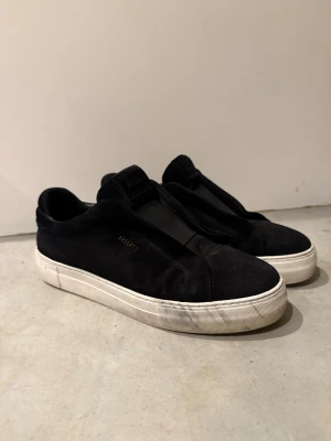 Axel Arigato - Svarta sneakers från Axel Arigato i mocka med vit platt sula och minimalistisk design. Skorna har elastiskt band över vristen istället för klassisk snörning och diskret guldfärgad logga på sidan. Perfekta för en clean och stilren look.
