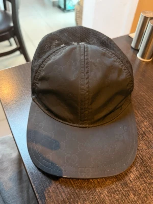 Svart meshkeps från Gucci - Säljer en svart keps från Gucci med diskret GG-mönster på framsidan och skärm. Baksidan är i mesh för extra ventilation och har en läderdetalj med Gucci-logga. Kepsen har en klassisk form och är tillverkad i syntetmaterial och läder.
